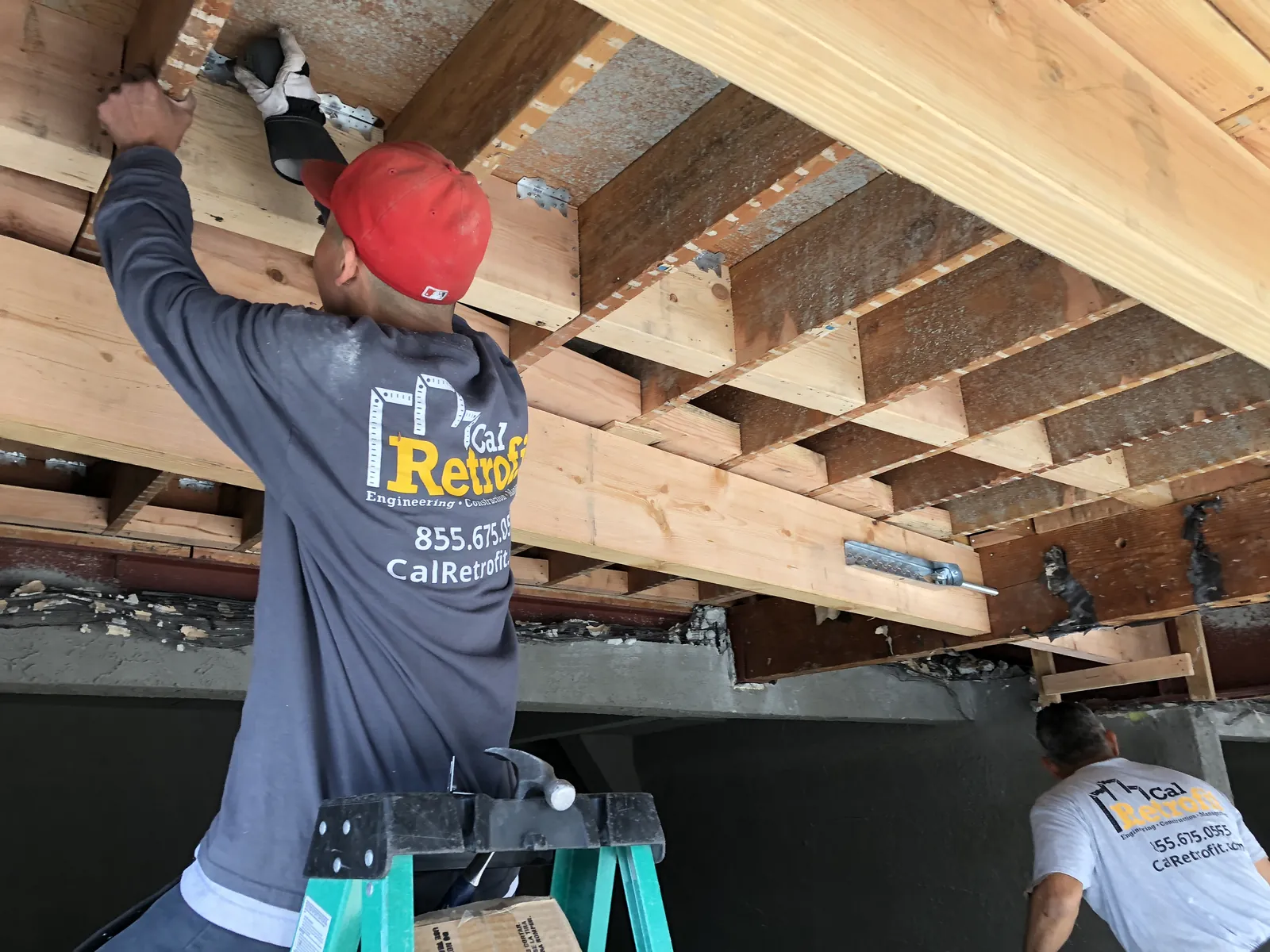 Cal Retrofit crew member fastening structural anchors to the underside of a building's floor framing during a retrofit.
