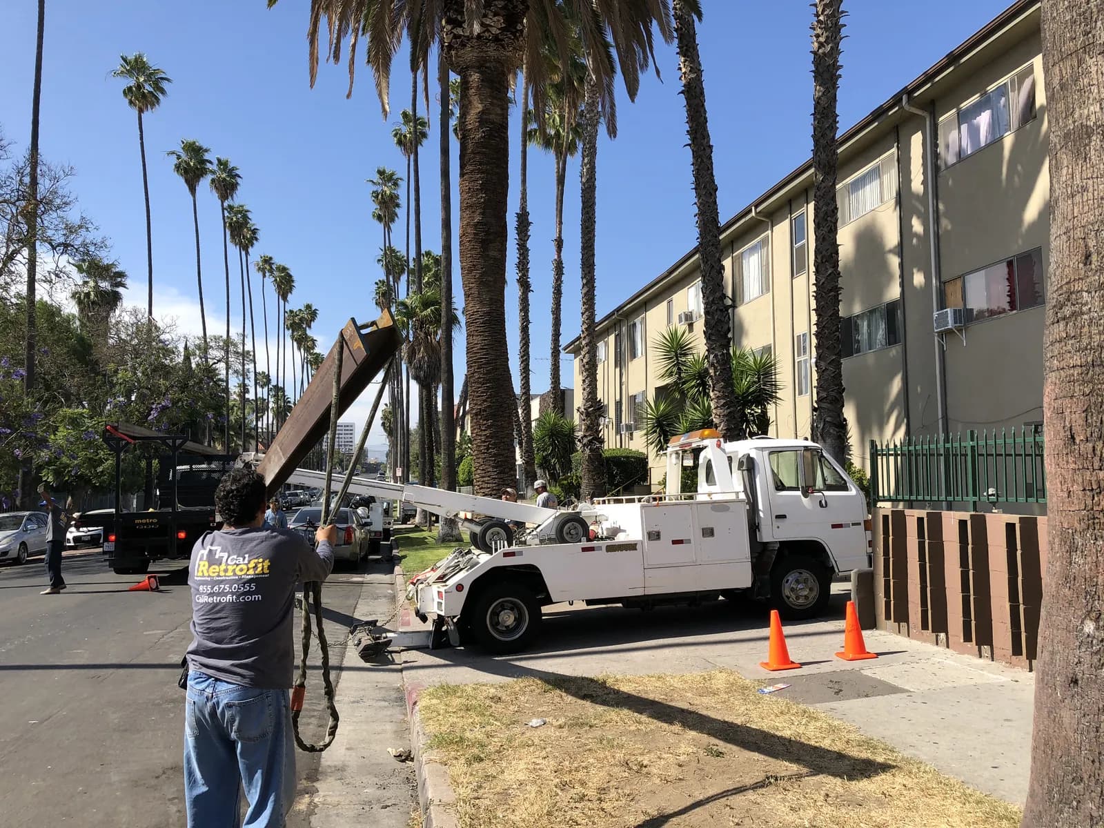 Cal Retrofit crew unloading a fabricated structural steel beam on a palm-lined Los Angeles street outside a multifamily retrofit job site.