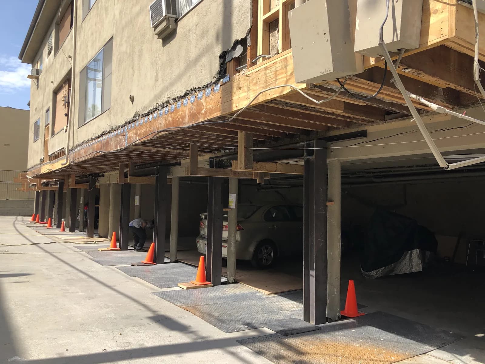 Row of temporary steel posts shoring a soft-story building while the permanent moment frames and footings are installed, with tenant parking still in use.
